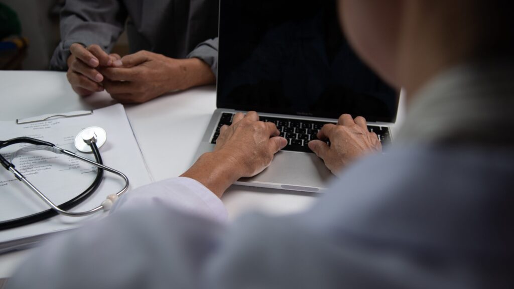 A doctor types on a laptop while a new NP sits across the desk with a clipboard and stethoscope nearby, discussing nurse practitioner tips.