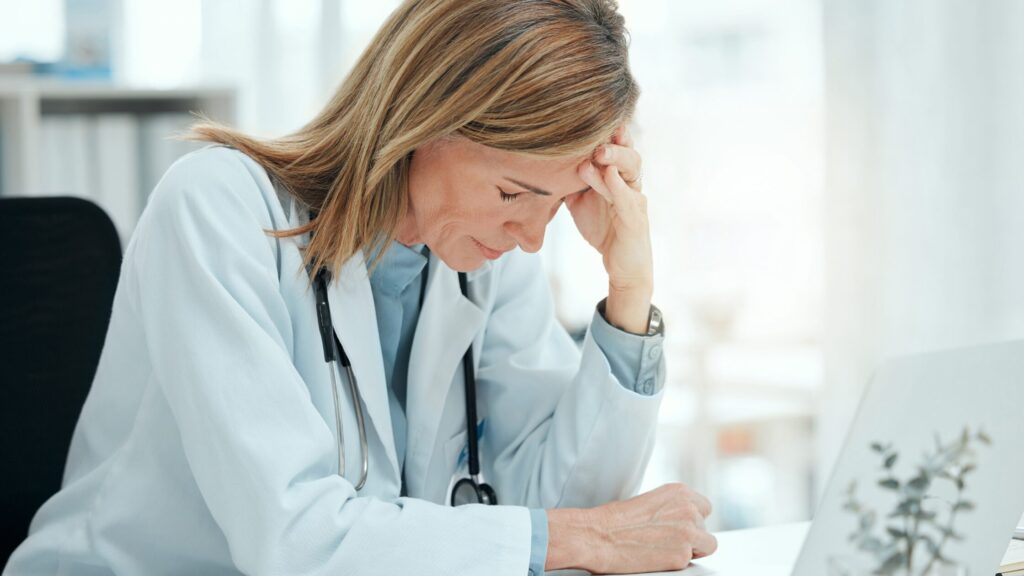 A female doctor sits at a desk with her head resting on her hand, looking down with a stressed expression—reflecting on how medical errors can become a valuable learning experience in medical education.