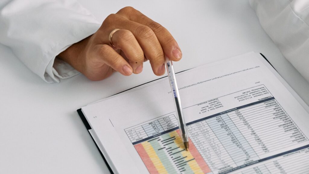 A hand holding a pen points at a highlighted row in a printed spreadsheet containing tables and color-coded data, illustrating the 7 steps to reduce medical errors for an improved learning experience.